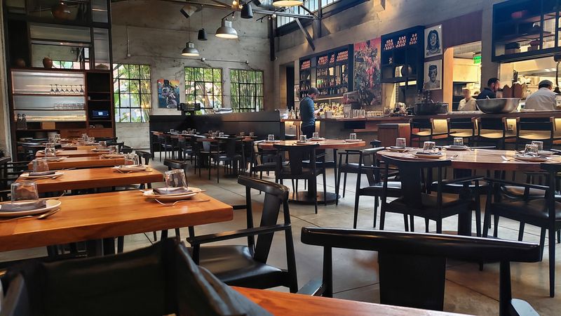 A restaurant dining room with long wooden tables set with upturned glasses and black chairs, under industrial pendant lamps, with a bar and open kitchen at the back and tall factory-style windows framing greenery.