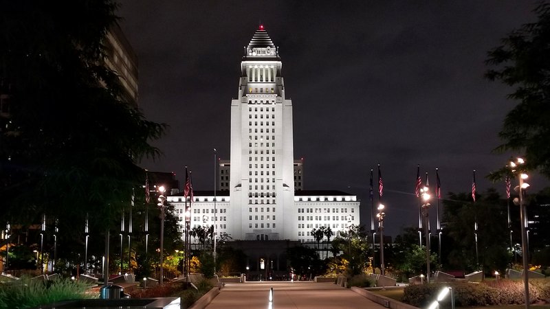 Los Angeles City Hall's white tower, lit against a dark night sky, seen from the end of a long illuminated walkway in Grand Park lined with flagpoles, lampposts and trees.